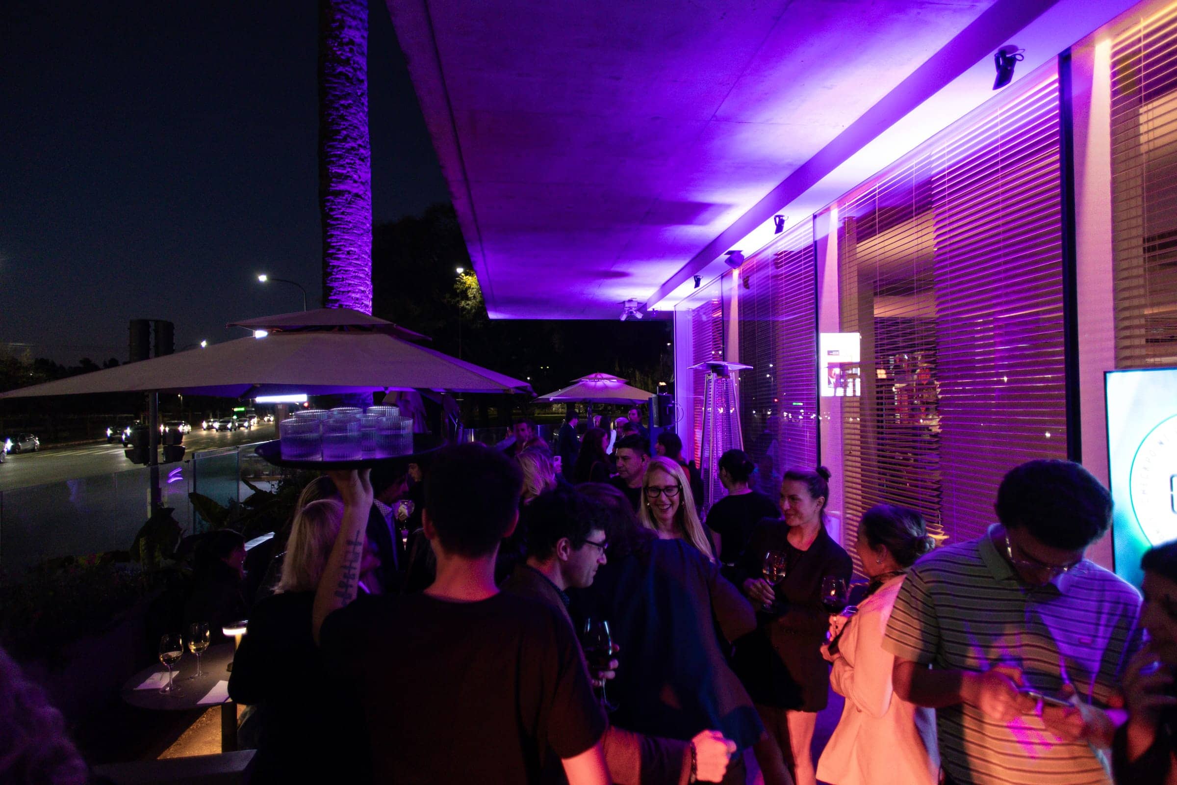 Cóctel de noche en la terraza, con invitados bajo las sombrillas y luz violeta sobre la fachada